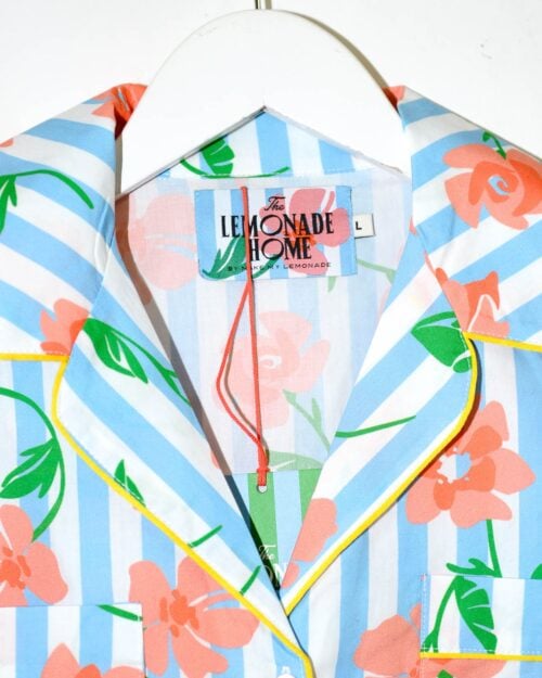 Kurzärmlige Bluse mit blau-weißen vertikalen Streifen und orangefarbenem Blumenmuster. Die Bluse hat gelbe Paspeln an Kragen und Knopfleiste, sowie zwei Brusttaschen. Sie hängt auf einem weißen Kleiderbügel.