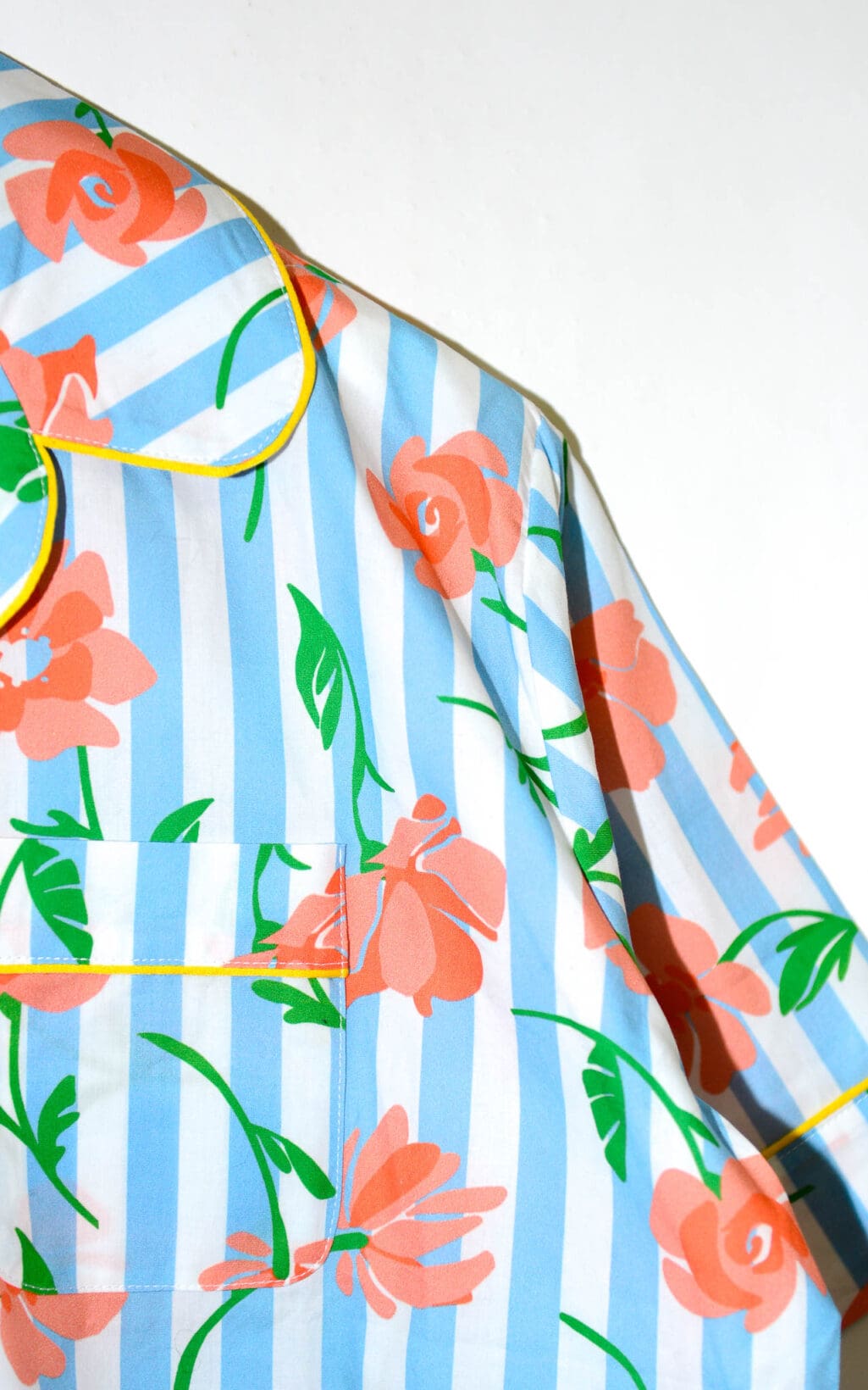 Kurz&auml;rmlige Bluse mit blau-wei&szlig;en vertikalen Streifen und orangefarbenem Blumenmuster. Die Bluse hat gelbe Paspeln an Kragen und Knopfleiste, sowie zwei Brusttaschen. Sie h&auml;ngt auf einem wei&szlig;en Kleiderb&uuml;gel.