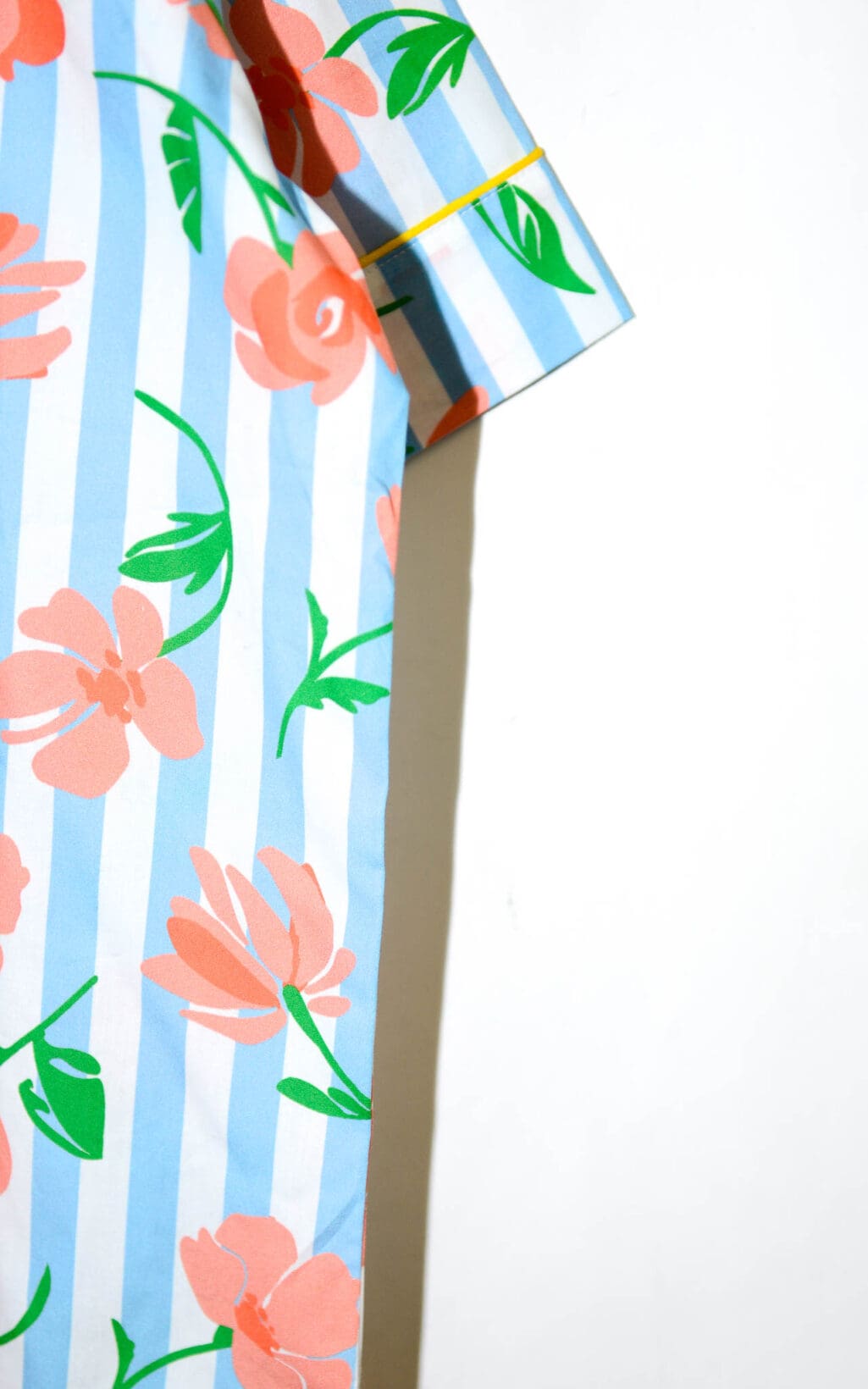 Kurz&auml;rmlige Bluse mit blau-wei&szlig;en vertikalen Streifen und orangefarbenem Blumenmuster. Die Bluse hat gelbe Paspeln an Kragen und Knopfleiste, sowie zwei Brusttaschen. Sie h&auml;ngt auf einem wei&szlig;en Kleiderb&uuml;gel.