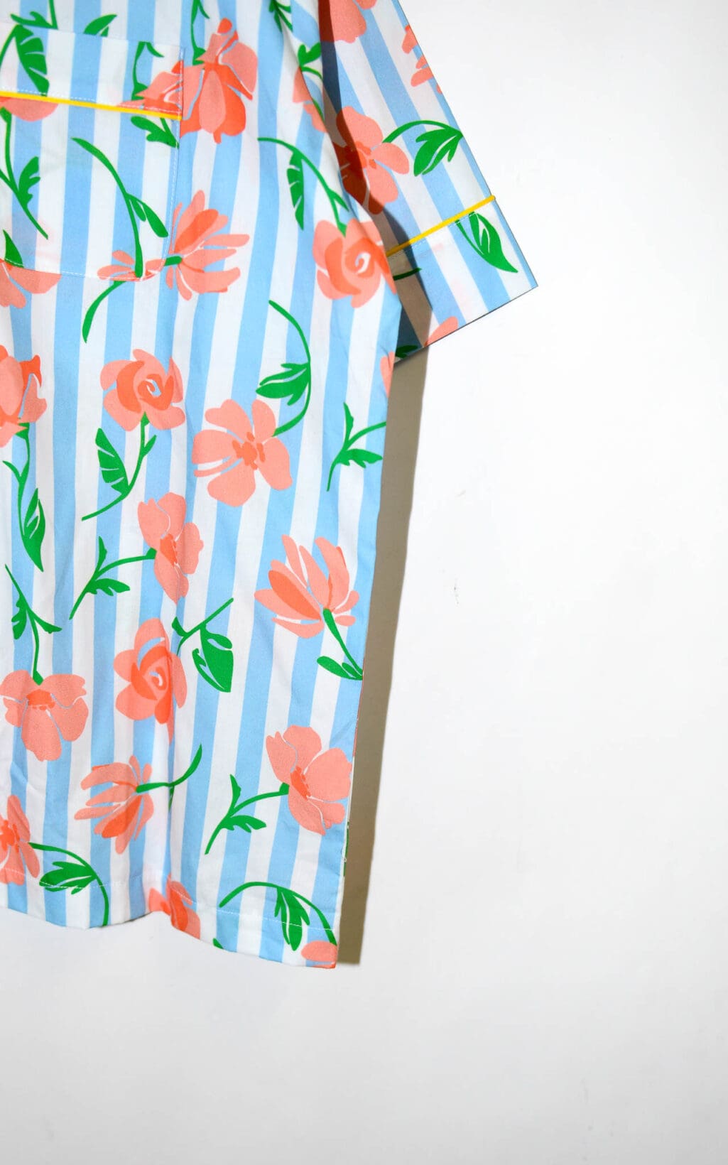 Kurz&auml;rmlige Bluse mit blau-wei&szlig;en vertikalen Streifen und orangefarbenem Blumenmuster. Die Bluse hat gelbe Paspeln an Kragen und Knopfleiste, sowie zwei Brusttaschen. Sie h&auml;ngt auf einem wei&szlig;en Kleiderb&uuml;gel.