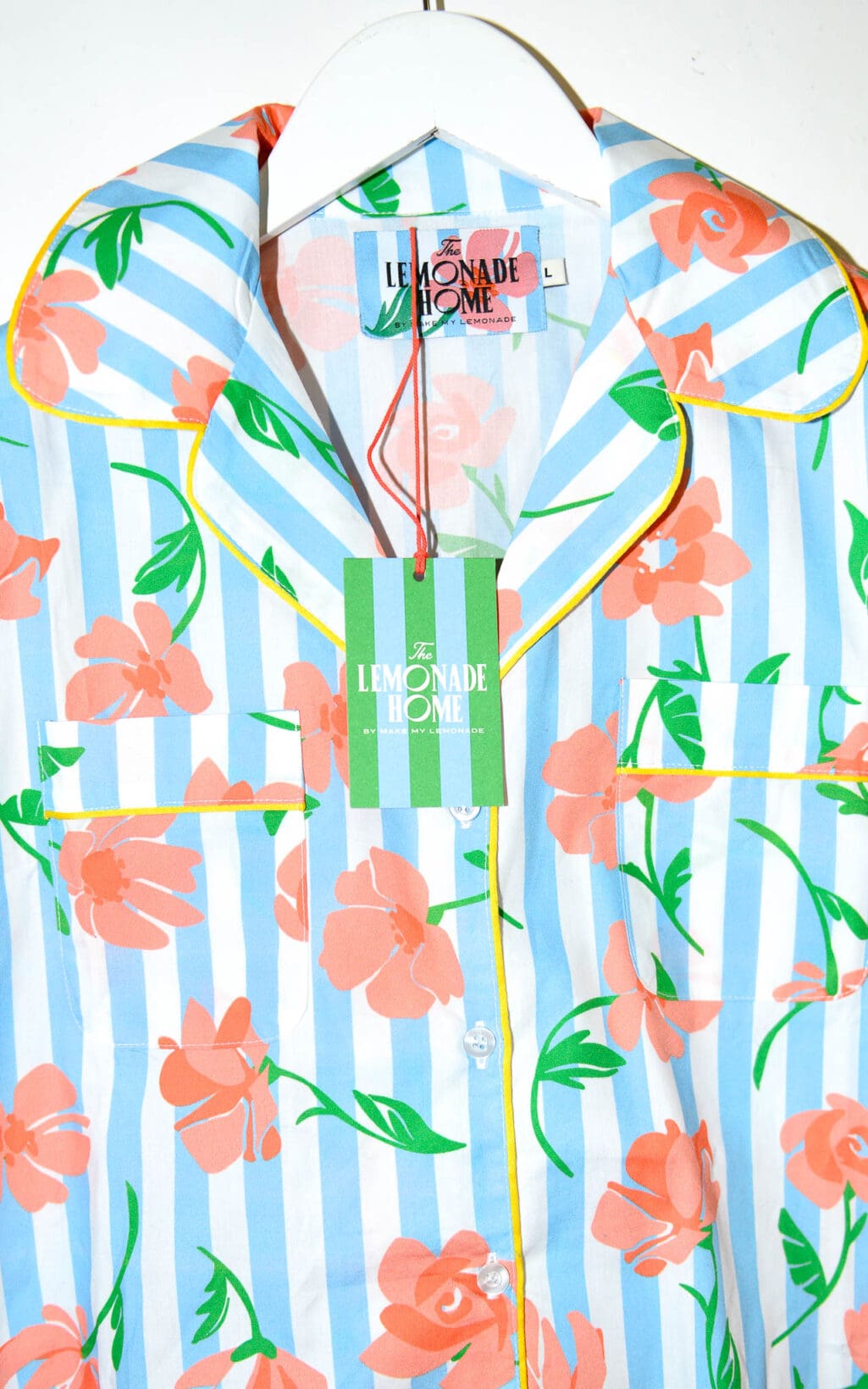 Kurz&auml;rmlige Bluse mit blau-wei&szlig;en vertikalen Streifen und orangefarbenem Blumenmuster. Die Bluse hat gelbe Paspeln an Kragen und Knopfleiste, sowie zwei Brusttaschen. Sie h&auml;ngt auf einem wei&szlig;en Kleiderb&uuml;gel.