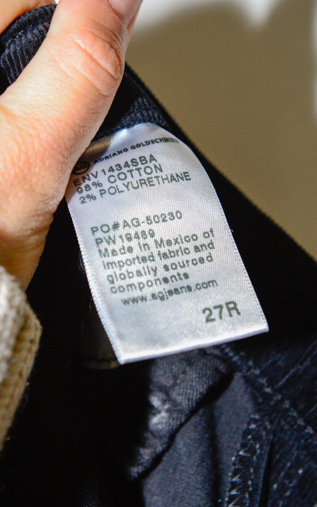 Eine Nahaufnahme eines zwischen den Fingern gehaltenen Kleidungsetiketts von Adriano Goldschmied Cigarette Leg &ndash; W27 zeigt die Stoffzusammensetzung (98 % Baumwolle, 2 % Polyurethan), Pflegehinweise und Herstellungsdetails auf dem Etikett.