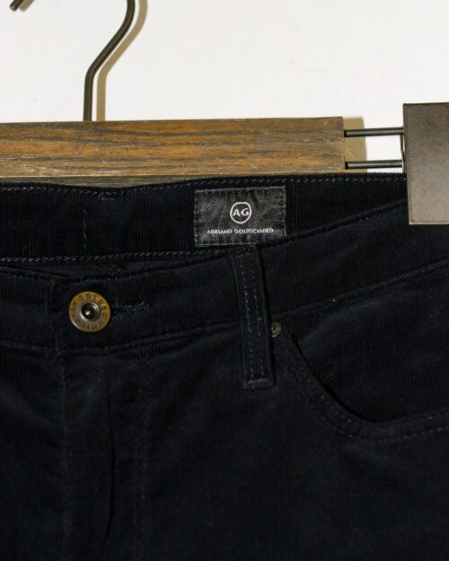 Nahaufnahme des Hosenbunds und der Vordertasche der dunkelblauen Cordhose Adriano Goldschmied Cigarette Leg &ndash; W27 auf einem Kleiderb&uuml;gel aus Holz, mit einem braunen Knopf und dem Etikett &bdquo;AG Adriano Goldschmied&ldquo; am inneren Hosenbund.