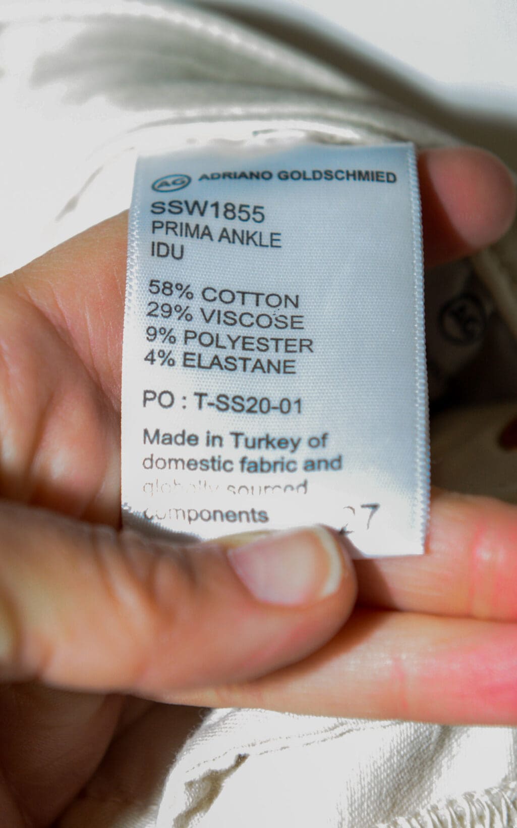 Eine Hand h&auml;lt ein Etikett von Adriano Goldschmied Prima Ankle Jeans &ndash; W27, auf dem der Stoffgehalt (58 % Baumwolle, 29 % Viskose, 9 % Polyester, 4 % Elastan) angegeben ist und das darauf hinweist, dass die Jeans in der T&uuml;rkei hergestellt wurde.