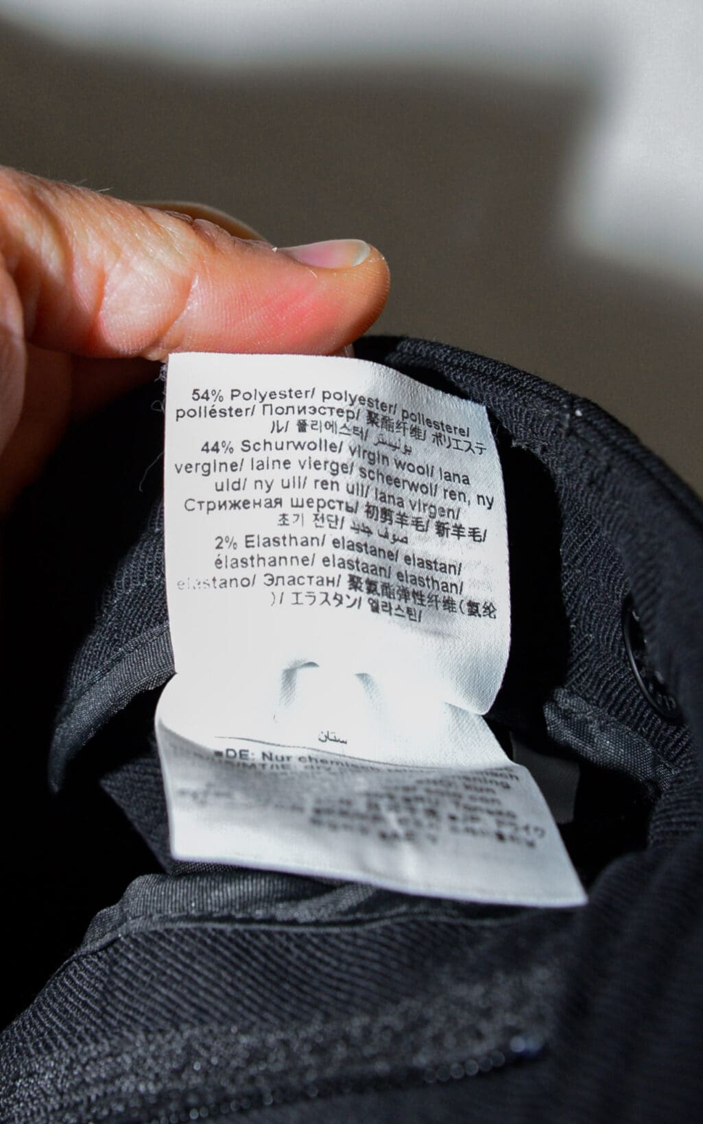 Eine Nahaufnahme einer Hand, die ein Kleidungsetikett der Closed Arlo Hose &ndash; W26 h&auml;lt. Die Stoffzusammensetzung ist in mehreren Sprachen angegeben: 54 % Polyester, 44 % Schurwolle und 2 % Elastan. Darunter ist schwarzer Stoff sichtbar.