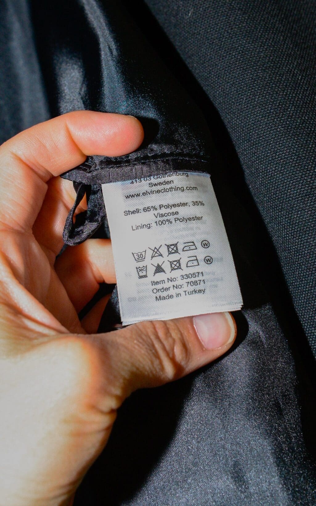 Eine Hand h&auml;lt das Etikett eines Elvine Blazers (Gr. M) auf schwarzem Stoff, auf dem Pflegesymbole, Materialien (Au&szlig;enmaterial 65 % Polyester/35 % Viskose; Futter 100 % Polyester) und &bdquo;Made in Turkey&ldquo; zu sehen sind.