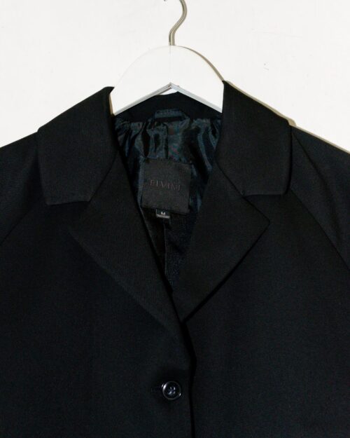 Der Elvine Blazer – Gr. M ist ein schwarzer Blazer mit Reverskragen, einem einzelnen sichtbaren Knopf und satinartigem Futter, der auf einem weißen Kleiderbügel vor einer weißen Wand hängt.