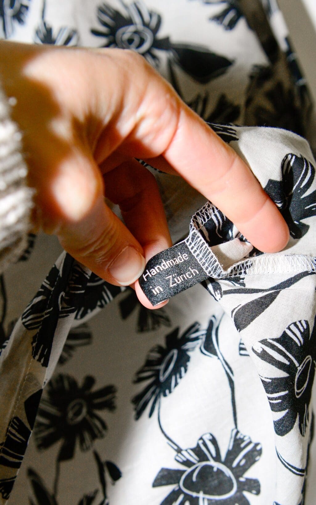 Eine Hand h&auml;lt den Rand des Sne Tops &ndash; Gr. L, einem wei&szlig;en Stoff mit schwarzen Blumendrucken, auf dem ein Etikett mit der Aufschrift &bdquo;Handmade in Z&uuml;rich&ldquo; zu sehen ist.