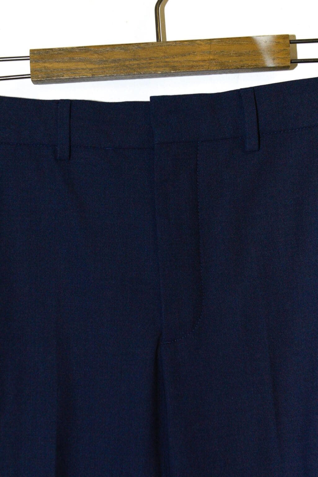 Eine Nahaufnahme des Hosenbunds und des oberen Teils der dunkelblauen Anzughose CLOSED Auckley Hose &ndash; W26, die auf einem h&ouml;lzernen Kleiderb&uuml;gel vor einem schlichten Hintergrund h&auml;ngt.
