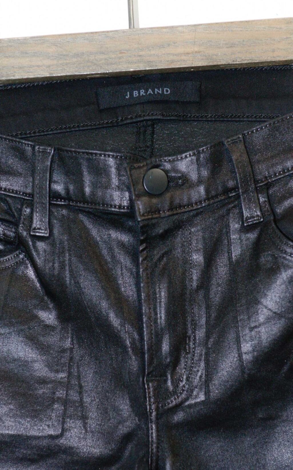 Schwarze beschichtete Jeans von J Brand in Lederoptik mit leichtem Schlag und klassischem 5-Pocket-Stil, auf einem Holzb&uuml;gel vor hellem Hintergrund pr&auml;sentiert.