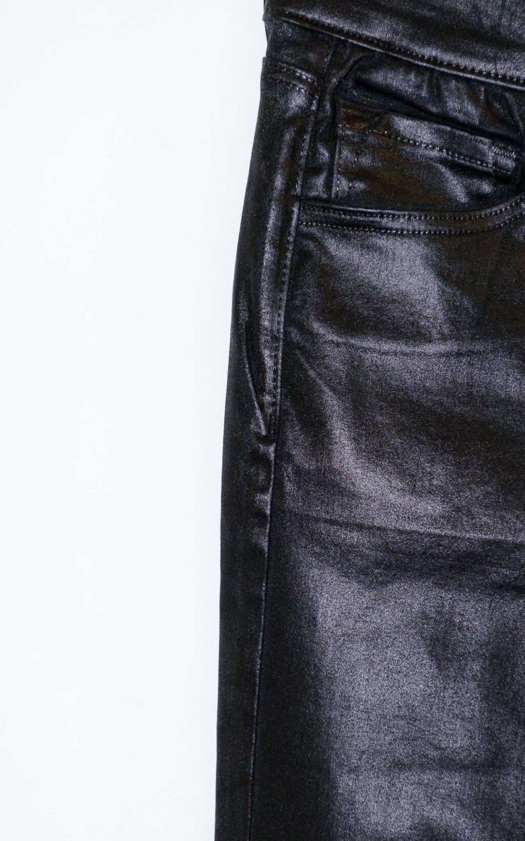 Schwarze beschichtete Jeans von J Brand in Lederoptik mit leichtem Schlag und klassischem 5-Pocket-Stil, auf einem Holzb&uuml;gel vor hellem Hintergrund pr&auml;sentiert.