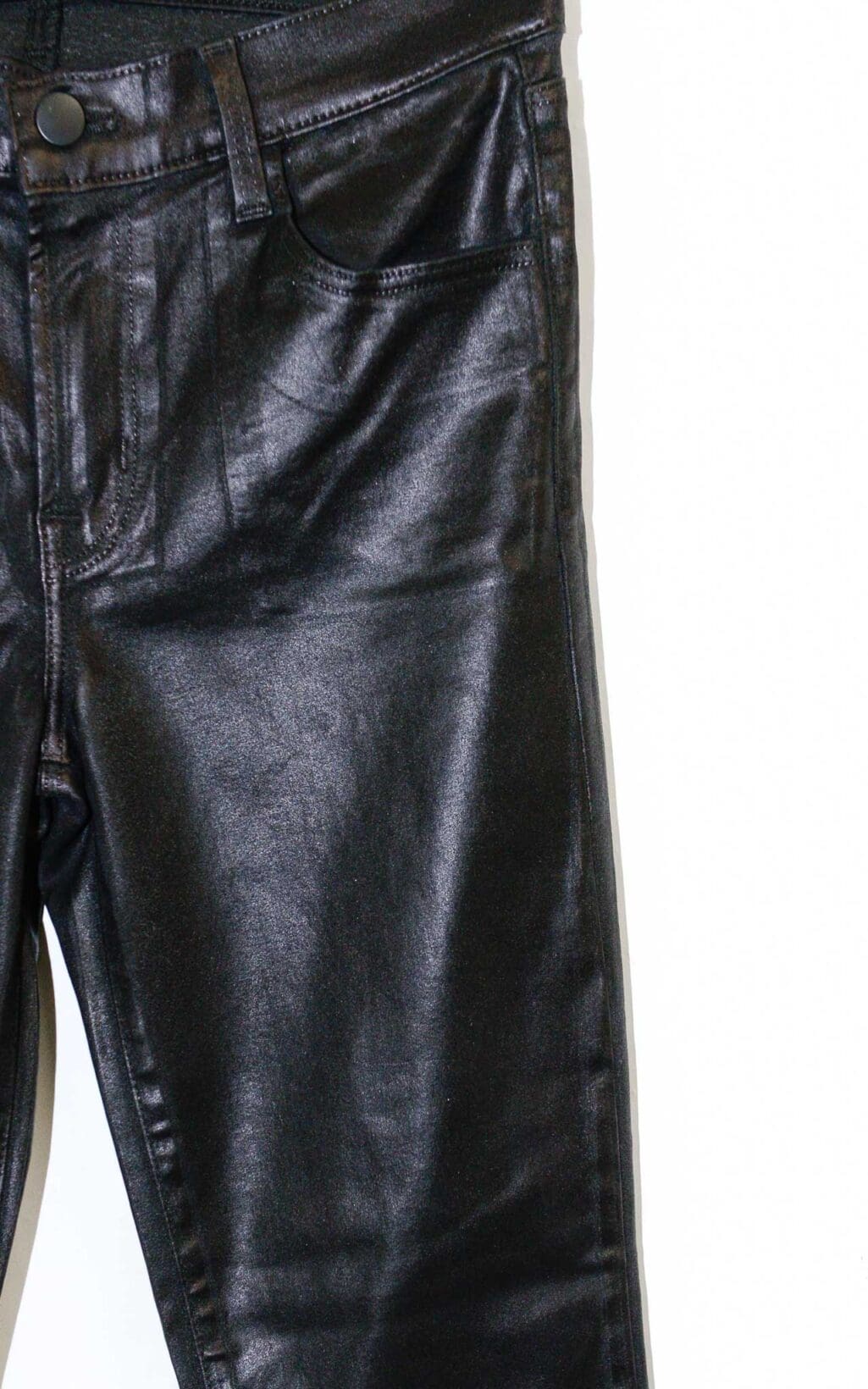 Schwarze beschichtete Jeans von J Brand in Lederoptik mit leichtem Schlag und klassischem 5-Pocket-Stil, auf einem Holzb&uuml;gel vor hellem Hintergrund pr&auml;sentiert.