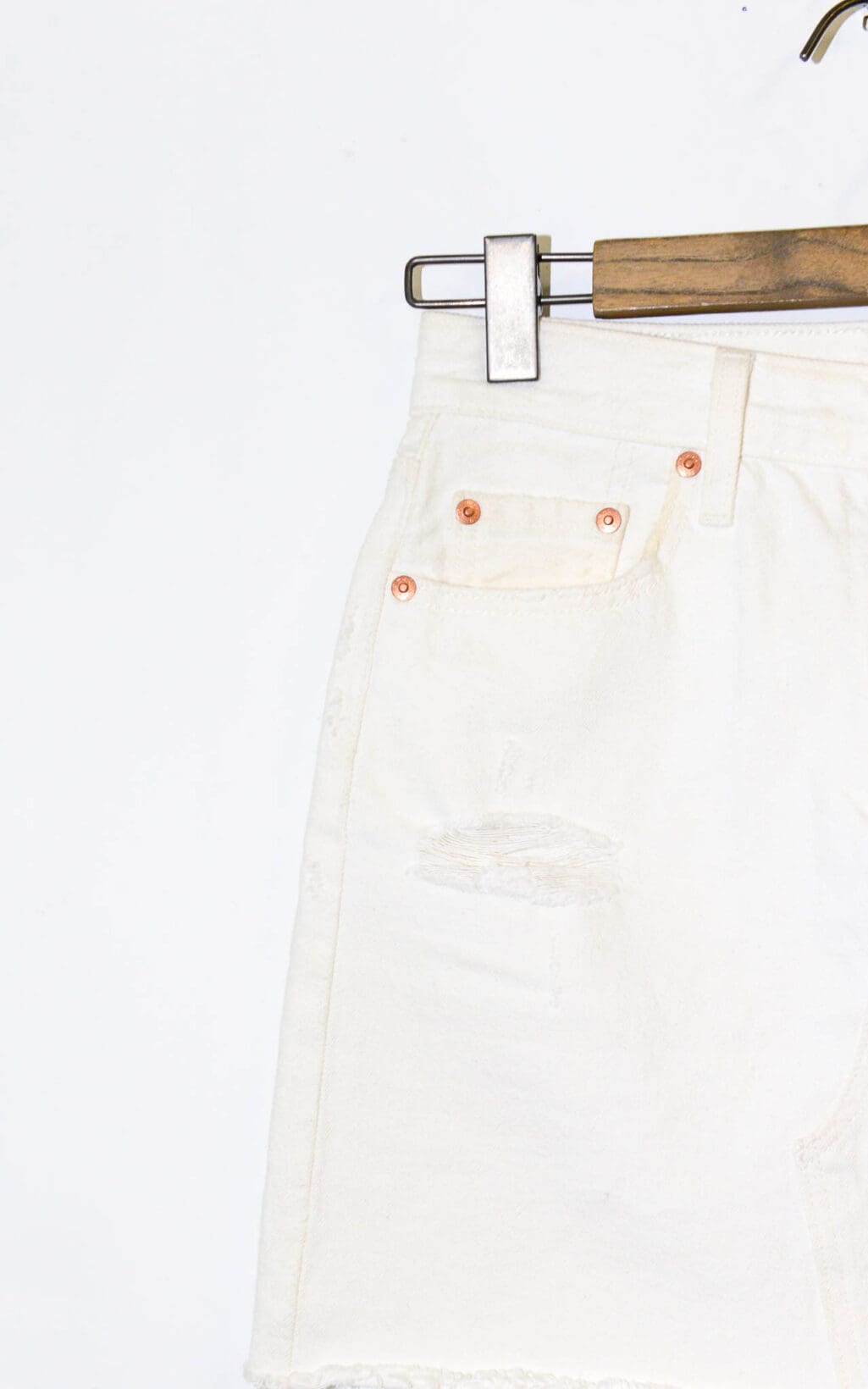 Nahaufnahme des Levi&lsquo;s Jeansrocks &ndash; W26, ein wei&szlig;er Jeansrock mit ausgefranstem Saum und kleinem Rissdetail unter der Tasche, der auf einem h&ouml;lzernen Kleiderb&uuml;gel vor einem schlichten wei&szlig;en Hintergrund h&auml;ngt.