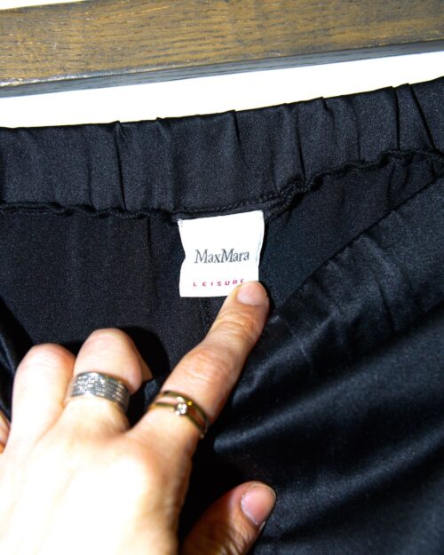 Eine Hand mit Ringen zeigt auf ein Kleidungsetikett im Inneren eines schwarzen Kleidungsst&uuml;cks mit der Aufschrift &bdquo;Maxmara Leisure Hose &ndash; Gr. S&ldquo;.