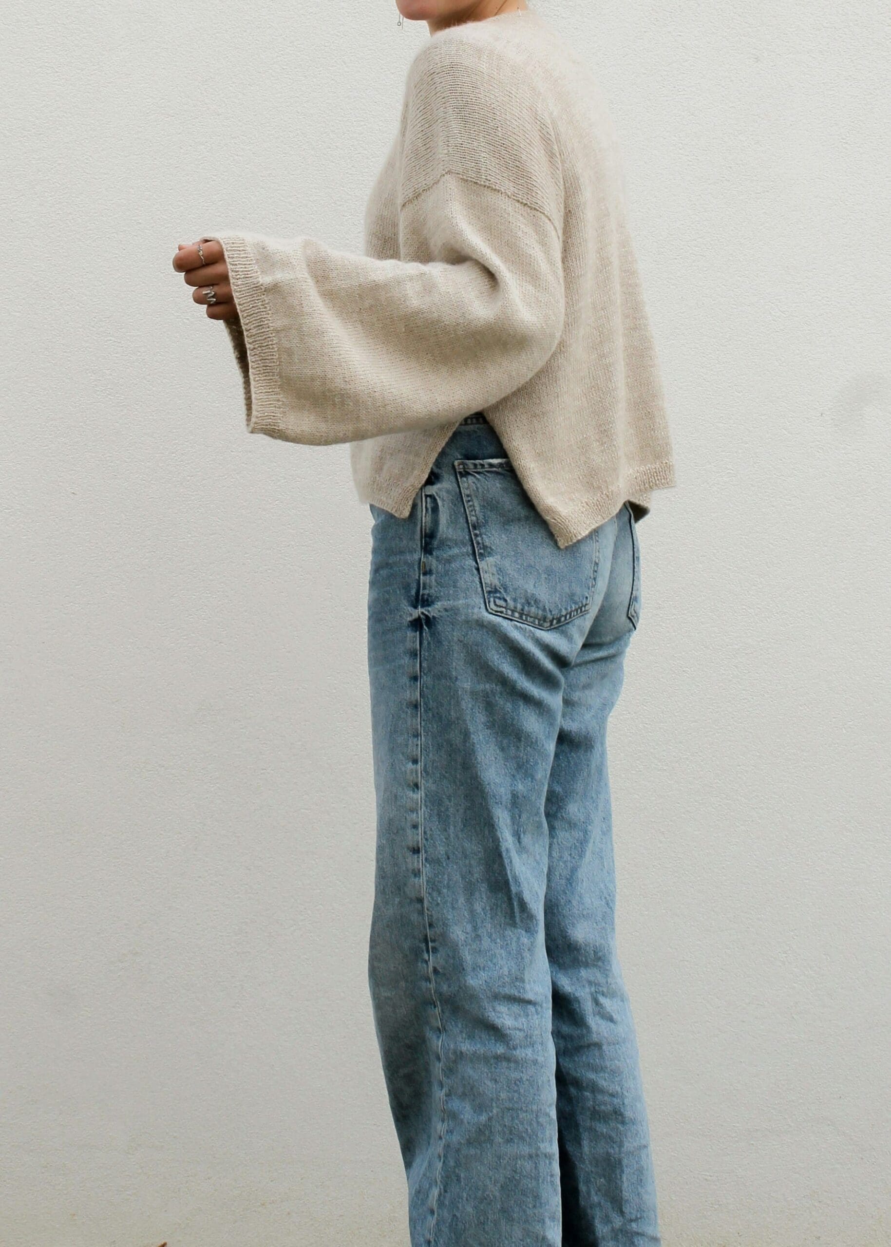 Eine Person sitzt auf einem Sofa. Sie trägt einen lockeren, hellbeigen Strickpullover und hellblaue Jeans. Ihre Hände liegen im Schoß, ihr Gesicht ist nicht zu sehen.
