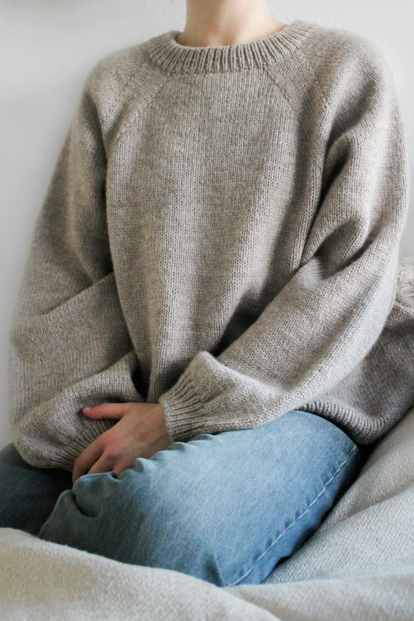 Eine Person sitzt auf einem Sofa. Sie trägt einen lockeren, hellbeigen Strickpullover und hellblaue Jeans. Ihre Hände liegen im Schoß, ihr Gesicht ist nicht zu sehen.