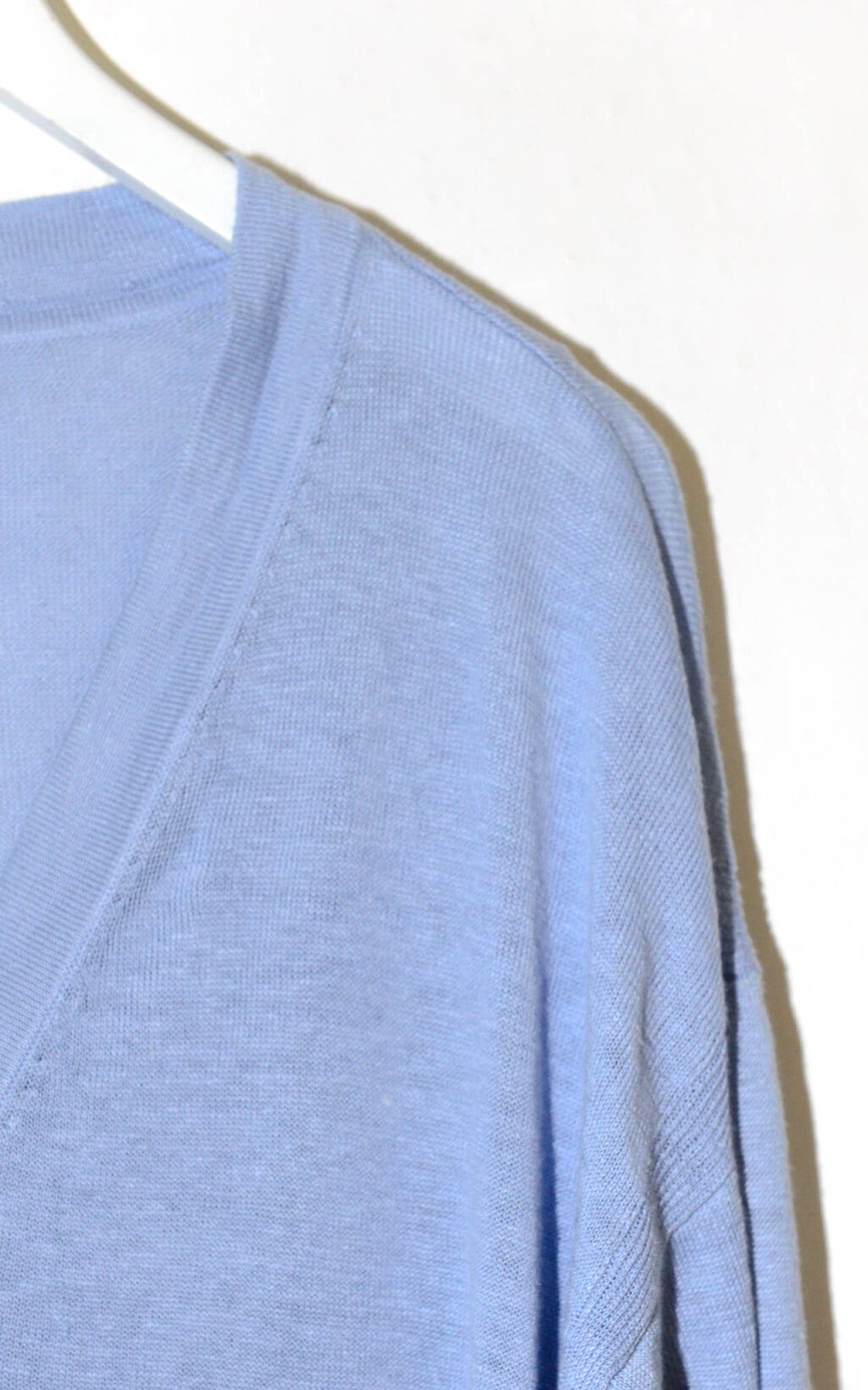 Ein Talbots Cardigan in Gr&ouml;&szlig;e L, hellblauer Strick, h&auml;ngt auf einem wei&szlig;en Kleiderb&uuml;gel vor einem schlichten wei&szlig;en Hintergrund, wobei das Sonnenlicht einen Schatten an die Wand wirft.