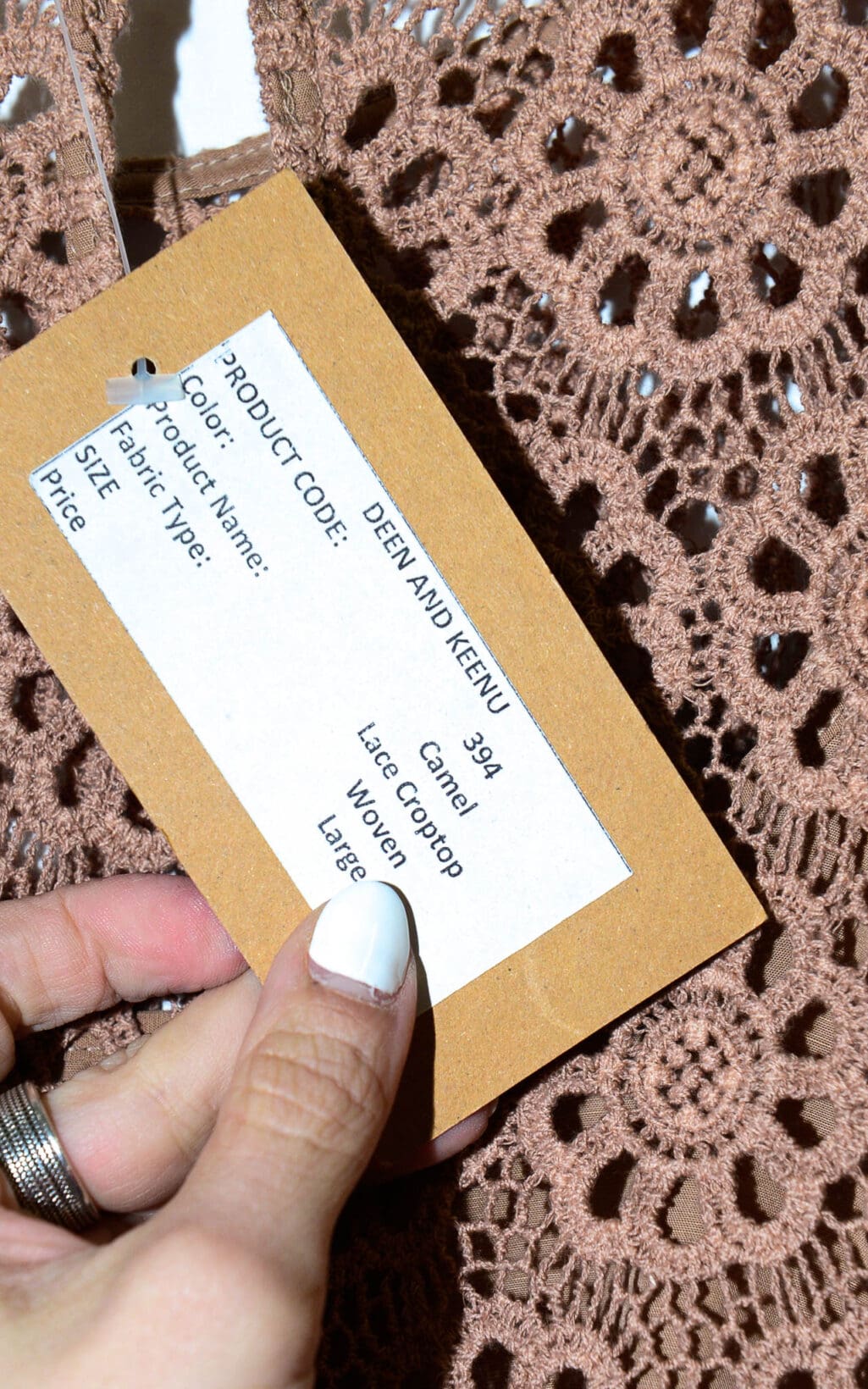 Eine Hand mit wei&szlig;em Nagellack h&auml;lt ein braunes Etikett mit der Aufschrift &bdquo;Deen & Keenu Lace Crop Top &ndash; Gr. L&ldquo;, das an einem kamelfarbenen, spitzengewebten Crop Top mit floralem H&auml;kelmuster befestigt ist. Auf dem Etikett sind Farbe, Produktname, Stoffart, Gr&ouml;&szlig;e und Preis aufgef&uuml;hrt.