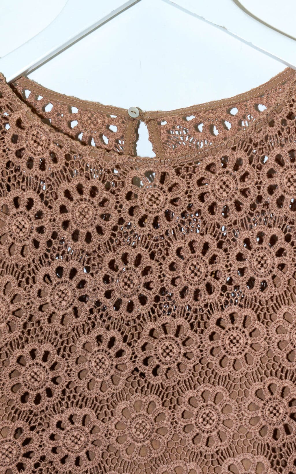 Nahaufnahme des Deen & Keenu Lace Crop Tops (Gr. L), einem braunen H&auml;keltop mit kreisf&ouml;rmigen Blumenmotiven, das auf einem wei&szlig;en Kleiderb&uuml;gel h&auml;ngt. Zu den Besonderheiten geh&ouml;rt ein kleiner Knopfverschluss am hinteren Ausschnitt.