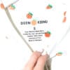 Eine Hand hält eine weiße Bluse von Deen & Keenu – Div. Grössen mit kleinen orangefarbenen Fruchtaufdrucken offen und gibt den Blick auf ein Etikett mit Pflegehinweisen und „Made in Pakistan“ frei.