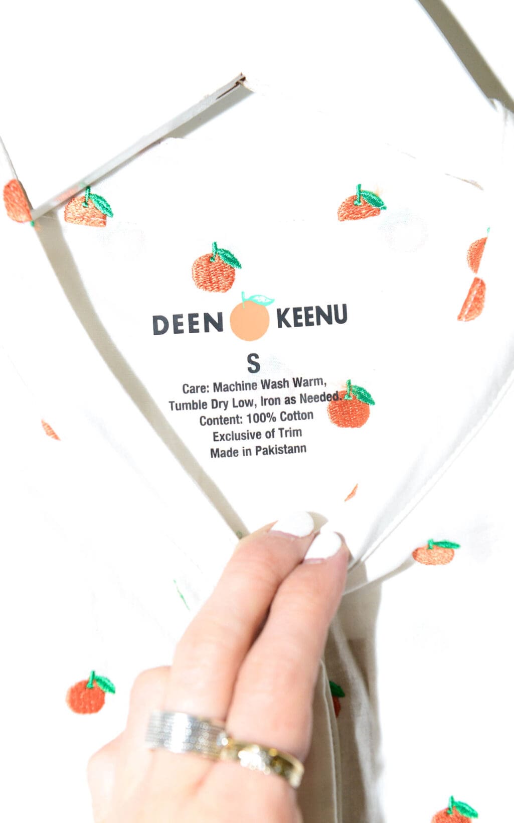 Eine Hand hält eine weiße Bluse von Deen & Keenu – Div. Grössen mit kleinen orangefarbenen Fruchtaufdrucken offen und gibt den Blick auf ein Etikett mit Pflegehinweisen und „Made in Pakistan“ frei.