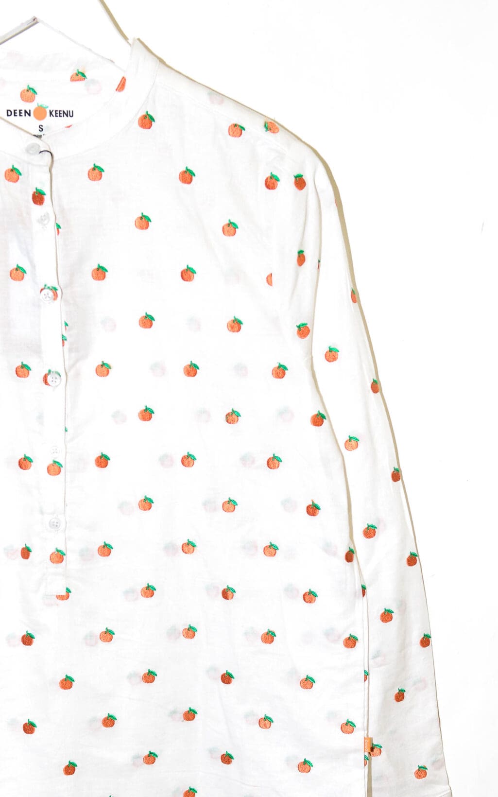 Die Deen & Keenu Bluse (verschiedene Größen) ist ein weißes Langarmshirt mit Stehkragen und kleinen orangefarbenen und grünen Fruchtaufdrucken, das auf einem weißen Kleiderbügel vor weißem Hintergrund hängt.