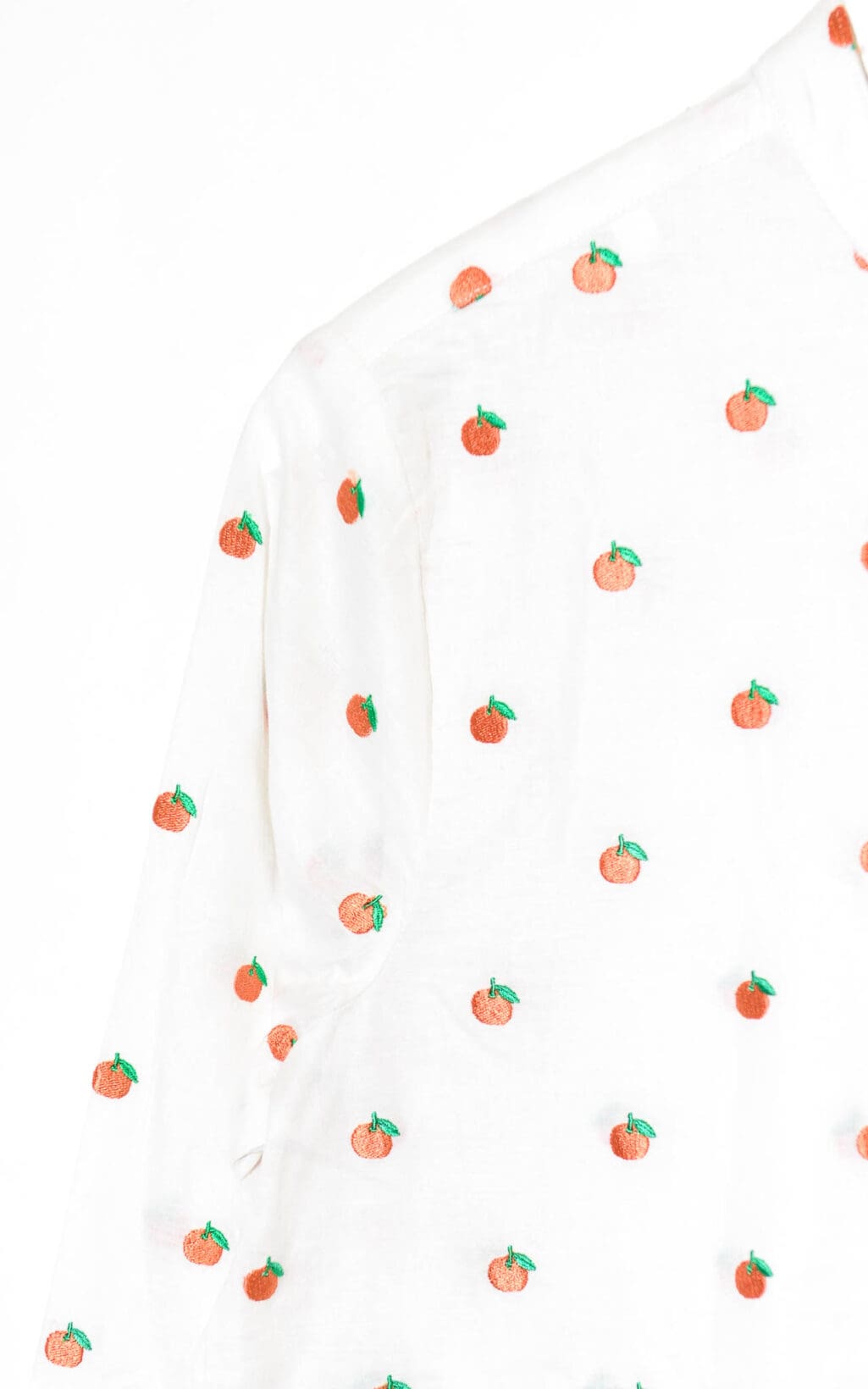 Eine Nahaufnahme der Bluse von Deen & Keenu (verschiedene Größen) zeigt weißen Stoff mit aufgestickten orangefarbenen und grünen Blättern, die Schulter und Ärmel hervorheben.