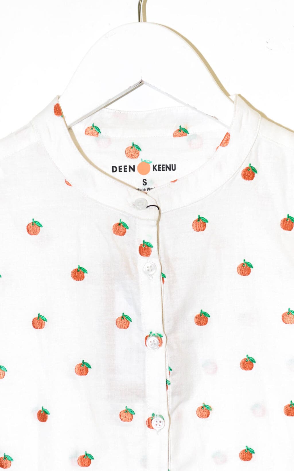 Ein weißes Hemd mit Stehkragen und einem sich wiederholenden orangefarbenen Fruchtaufdruck. Das Hemd mit der Aufschrift „Deen & Keenu Bluse – Div. Grössen“ hängt auf einem weißen Kleiderbügel.