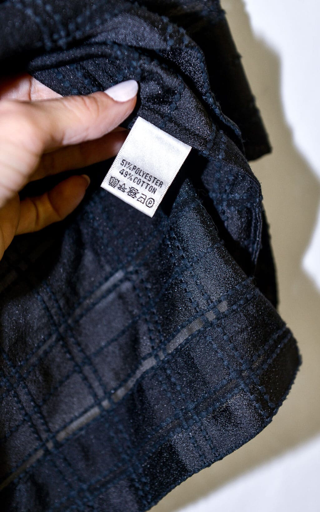 Eine Hand h&auml;lt die Globus Bluse &bdquo;Doha&ldquo; - Gr. 40 in schwarz kariert, darauf ist ein wei&szlig;es Etikett mit der Aufschrift &bdquo;51% POLYESTER, 49% BAUMWOLLE&ldquo; und Wasch- und Pflegesymbolen zu sehen.