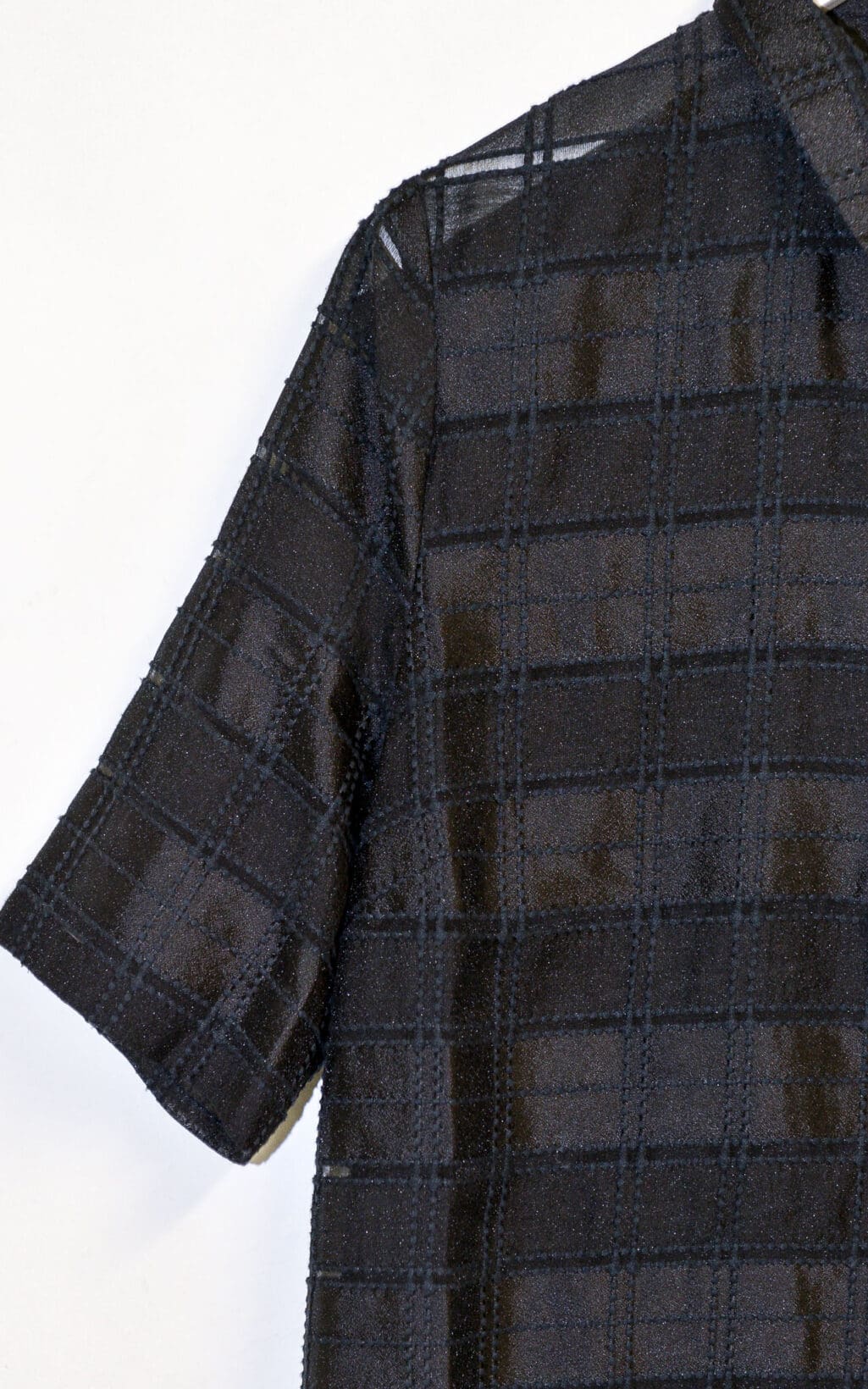 Nahaufnahme der Globus &bdquo;Doha&ldquo; Bluse (Gr. 40), einem transparenten schwarzen Kurzarmoberteil mit Karomuster, dessen strukturierter Stoff und N&auml;hte sich vor einem schlichten wei&szlig;en Hintergrund abheben.