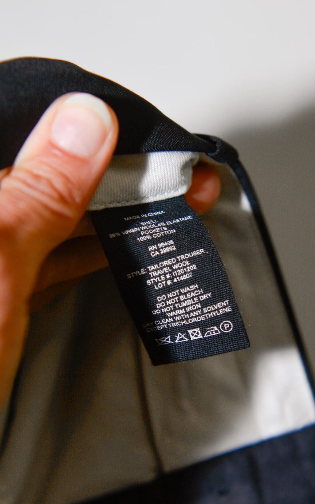 Eine Hand h&auml;lt ein schwarzes Kleidungsetikett hoch, das an der Theory Hose &ndash; Gr. 36 befestigt ist und Pflegehinweise, Materialzusammensetzung sowie Symbole zum Waschen, Trocknen und B&uuml;geln anzeigt.