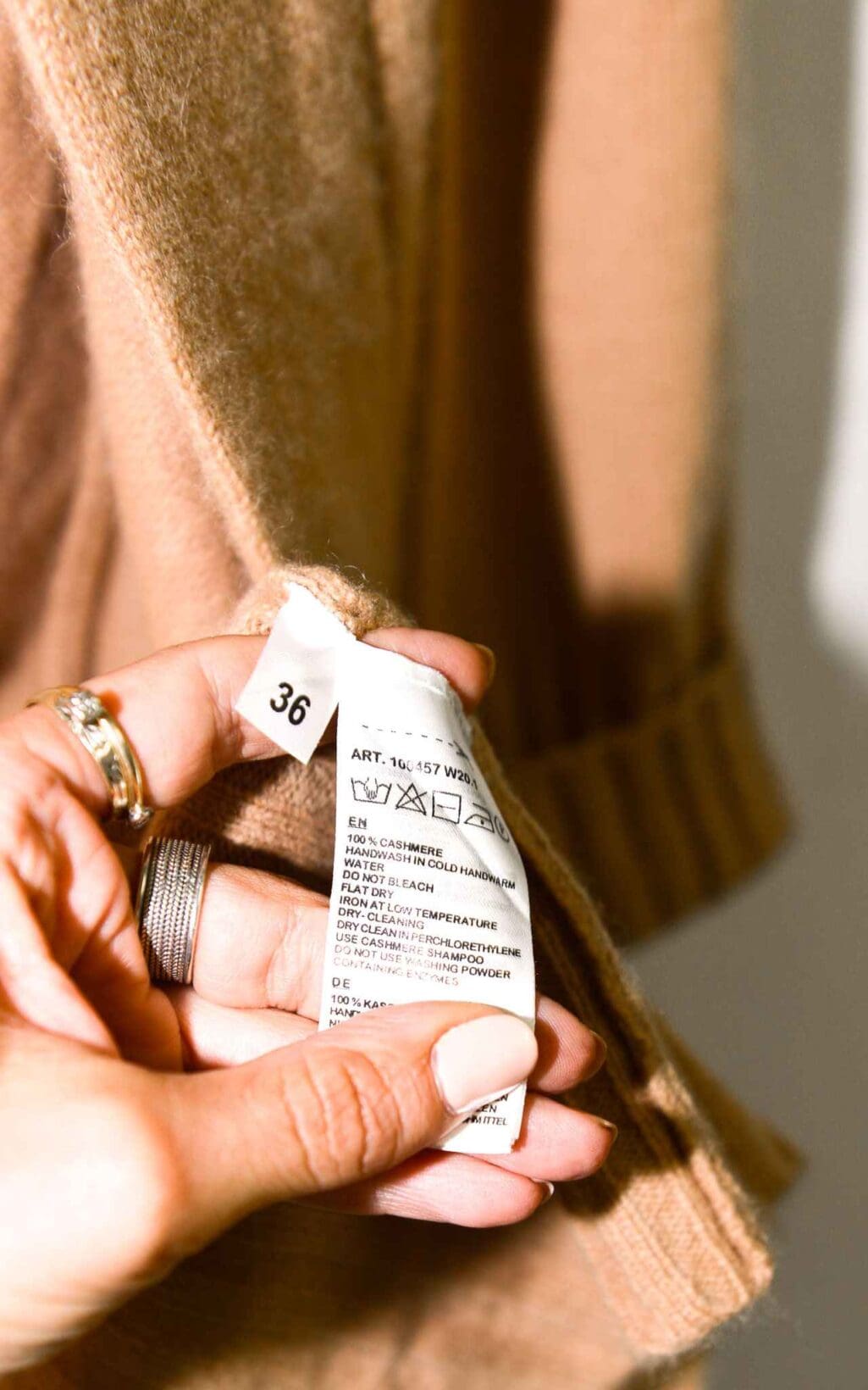 Eine Hand mit manik&uuml;rten N&auml;geln und silbernen Ringen h&auml;lt das Pflege- und Gr&ouml;&szlig;enetikett eines braunen Repeat Luxury Cardigan - Gr. 36, auf dem Waschhinweise und die Gr&ouml;&szlig;e "36" stehen.