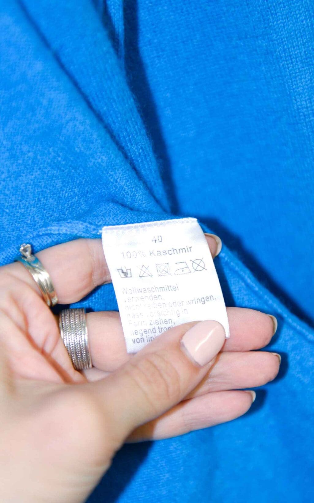 Eine Hand mit Ringen h&auml;lt das Kleidungsetikett einer Steffen Schraut 100% Kaschmir Strickjacke (Gr. 40) auf blauem Stoff. Das Etikett zeigt die Gr&ouml;&szlig;e 40, den Stoffgehalt als 100% Kaschmir und die Pflegehinweise auf Deutsch.